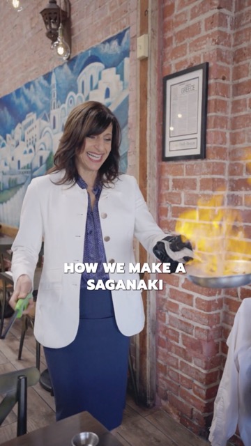 Nicky cooking saganaki with flames rising from the pan; on-image words introduce how it is made.